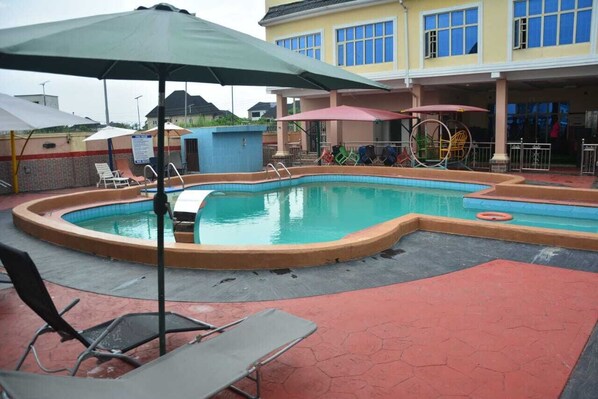 A swimming pool with lounge chairs and umbrellas, a covered area with a play structure, and a building with multiple windows.