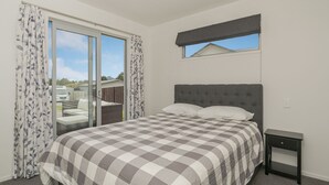 A bedroom with a bed, a nightstand, a window with a view, and a sliding door to a balcony. - A Sunshine Retreat - Pauanui Holiday Home (Pauanui)