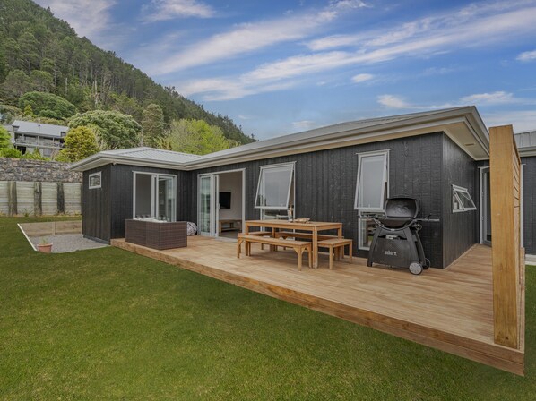 Een modern huis met een terras, een barbecue en een groot raam met uitzicht op een bebost heuvel.'