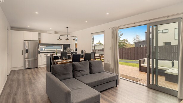 A modern living room with a grey sectional sofa, a kitchen with stainless steel appliances, and a sliding glass door leading to a patio with outdoor furniture. - A Sunshine Retreat - Pauanui Holiday Home (Pauanui)