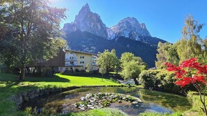 A mountain landscape with a building, a pond with lily pads, and trees. - FLORIS GREEN SUITES (Castelrotto)