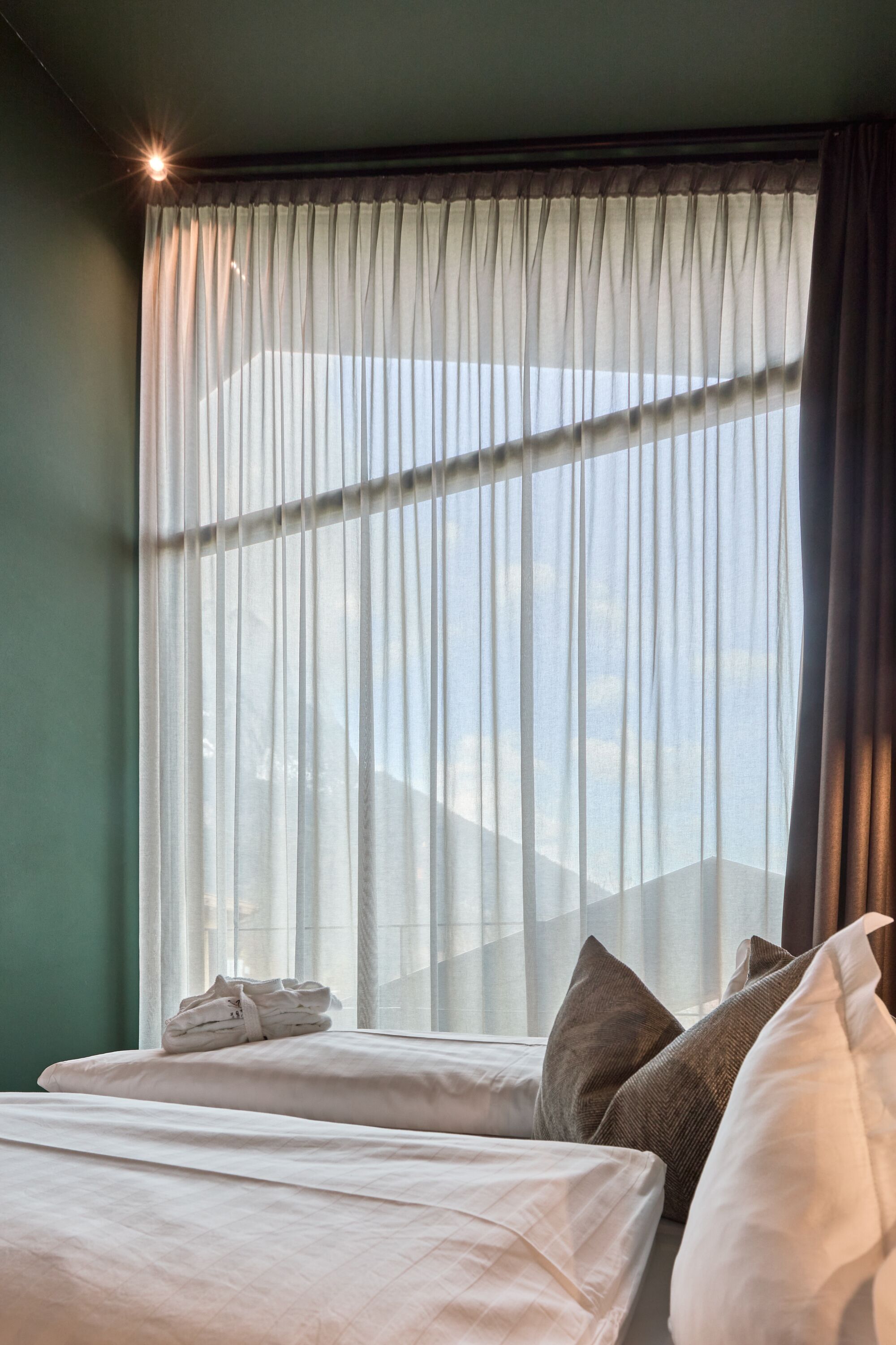 A hotel room with a bed, pillows, and a window with sheer curtains.
