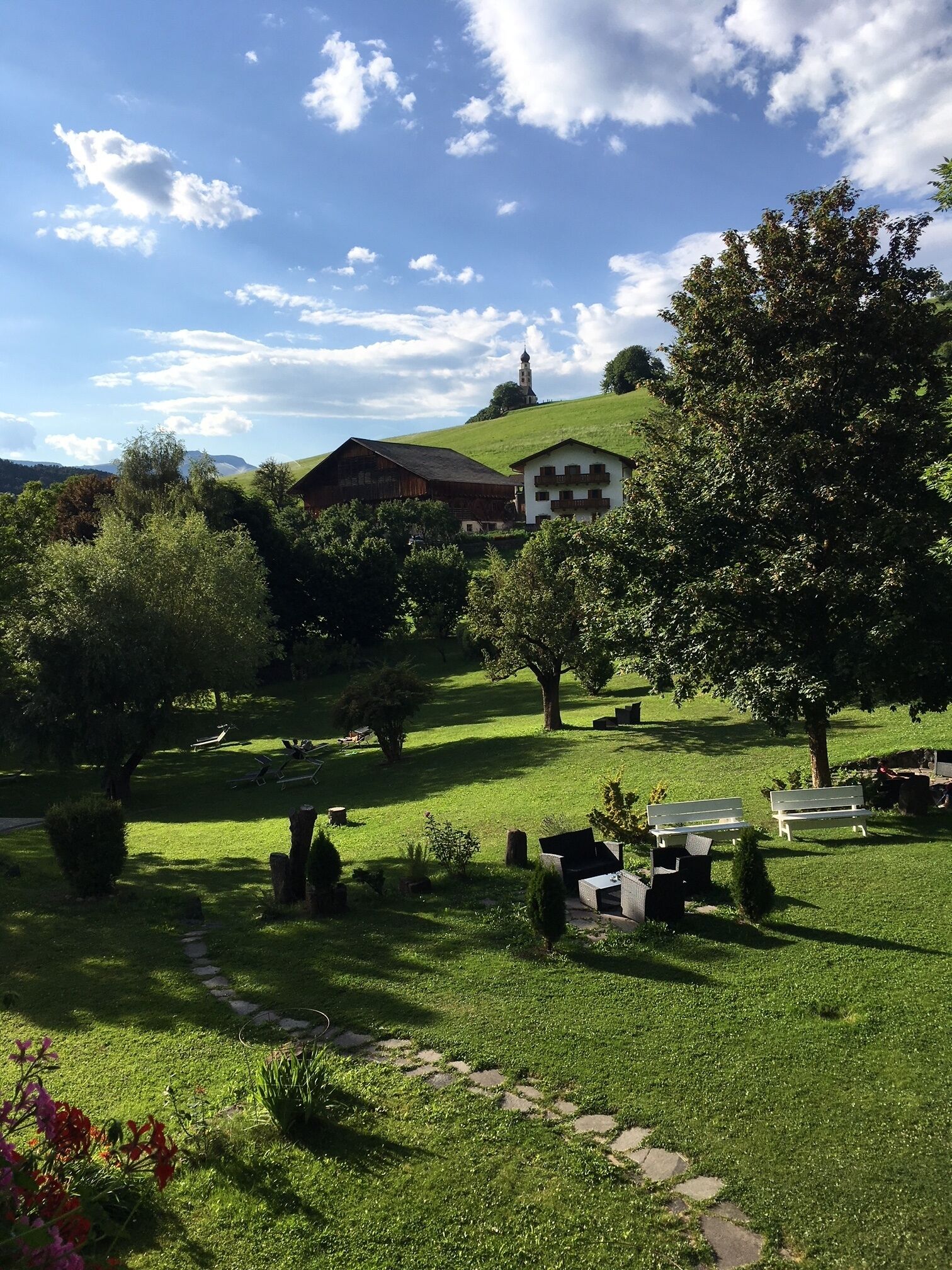 A rural landscape with a large barn, a white building with balconies, and a church on a hill.