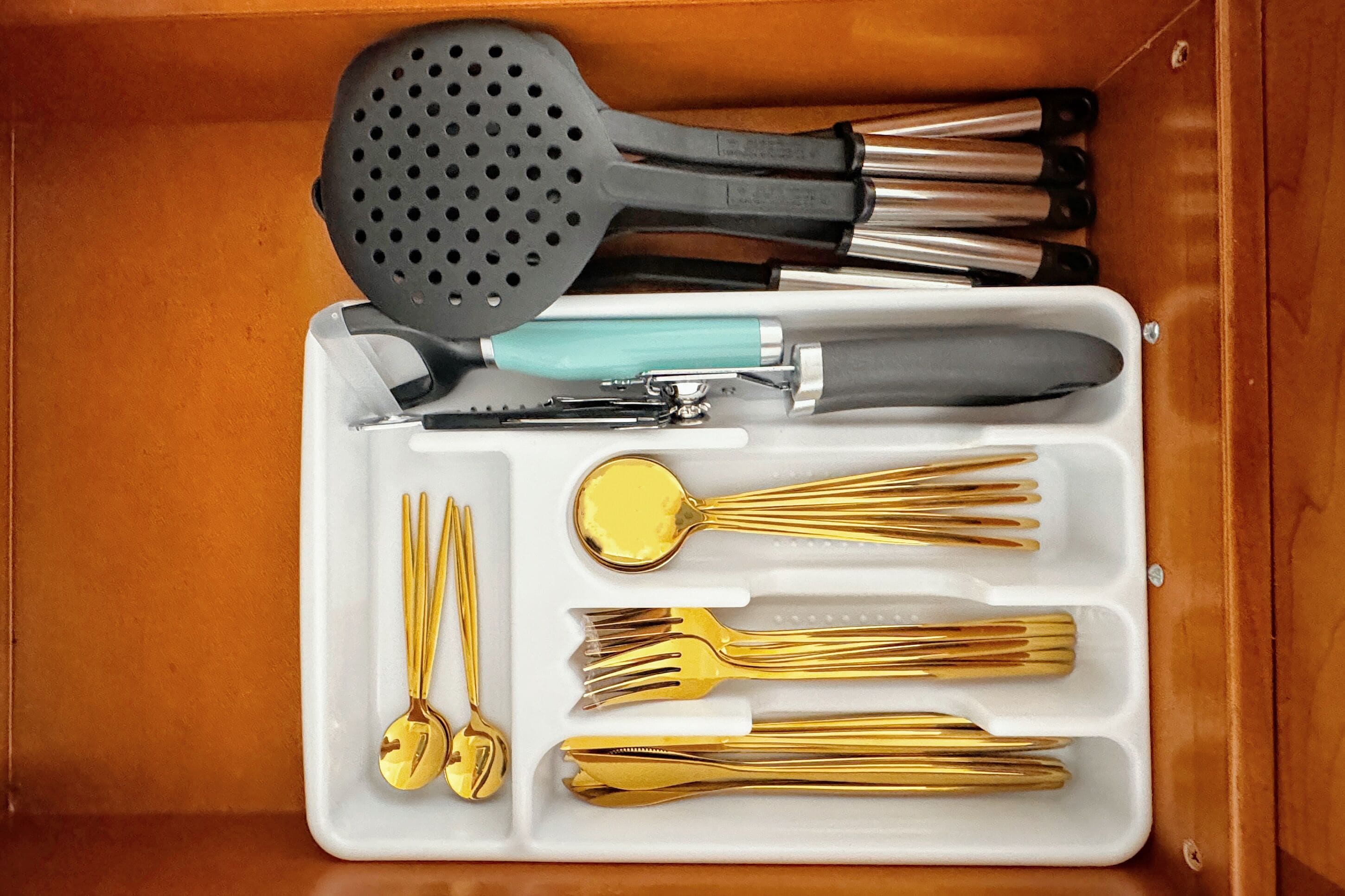 Un cassetto pieno di vari utensili da cucina, tra cui una spatola nera, un coltello con manico color acquamarina e diverse forchette e cucchiai dorati.