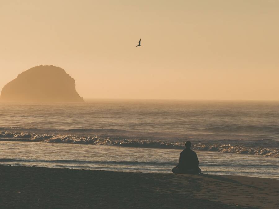 A beach scene with a person sitting on the sand, waves approaching, and two large rock formations in the distance.
