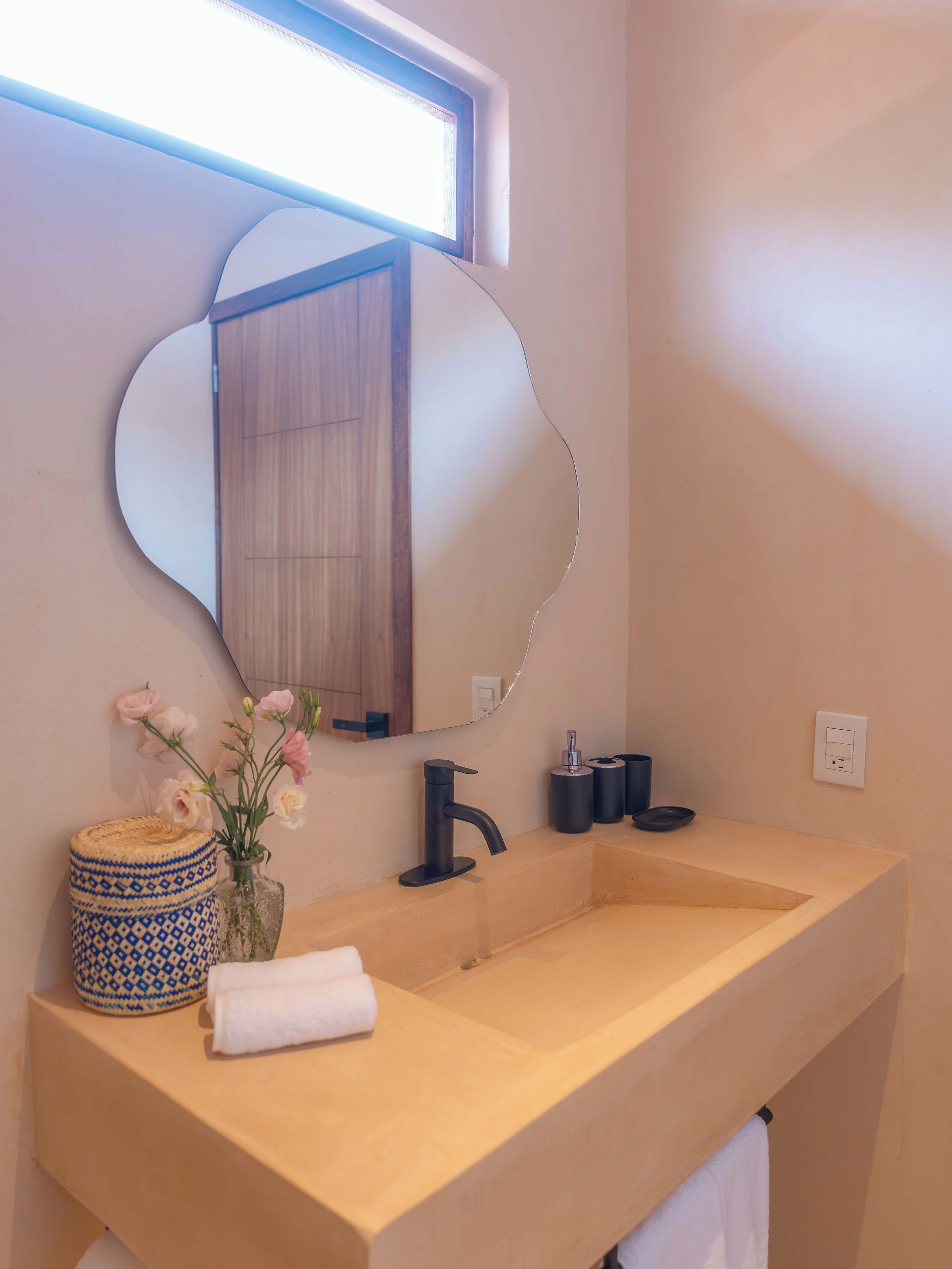Ein Badezimmer mit einem großen Spiegel, einem beige farbenen Waschbecken und einer Vase mit Blumen.