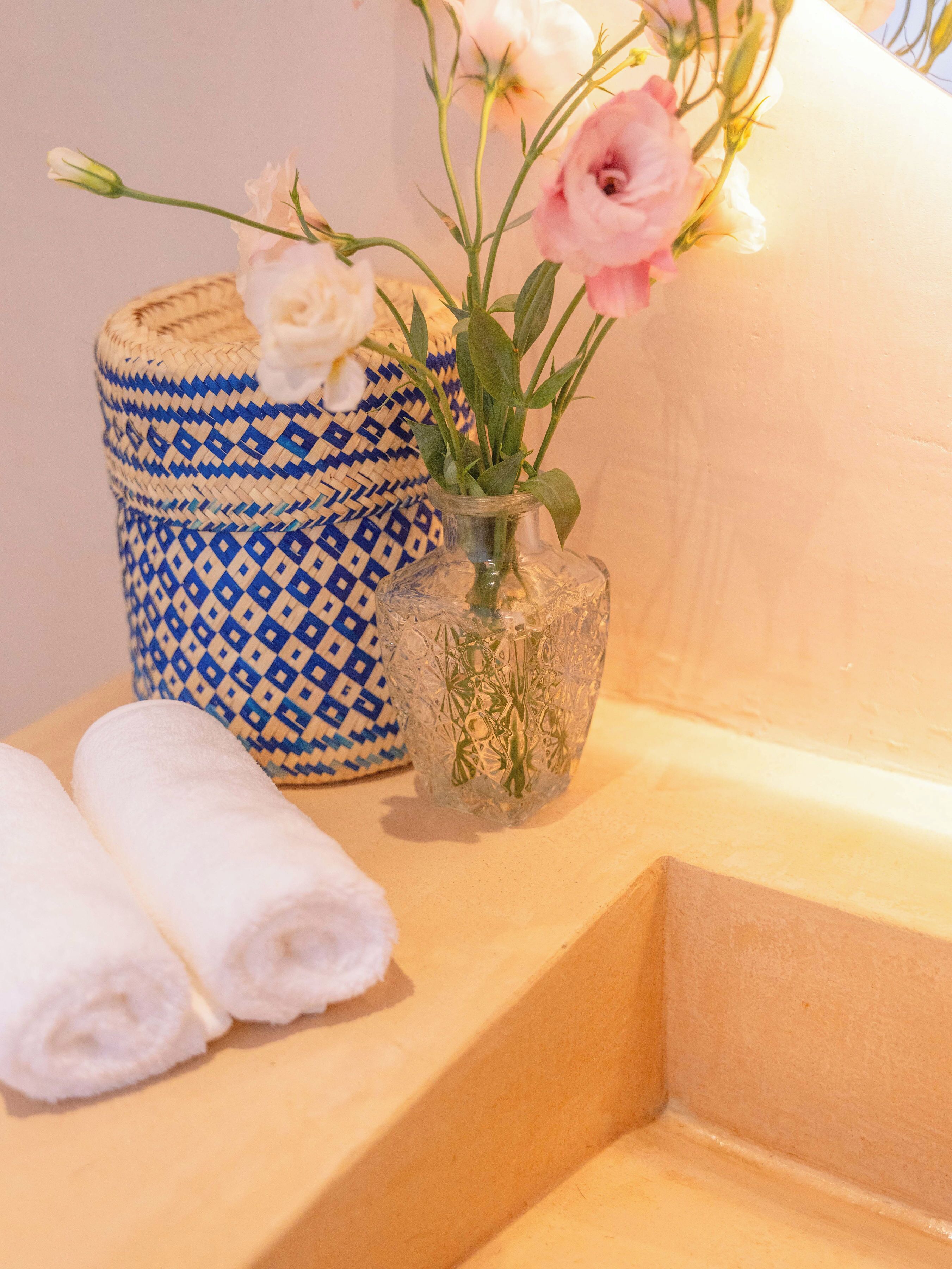 A bathroom with a blue and white patterned container, a clear glass vase with flowers, and neatly rolled towels on a beige surface.