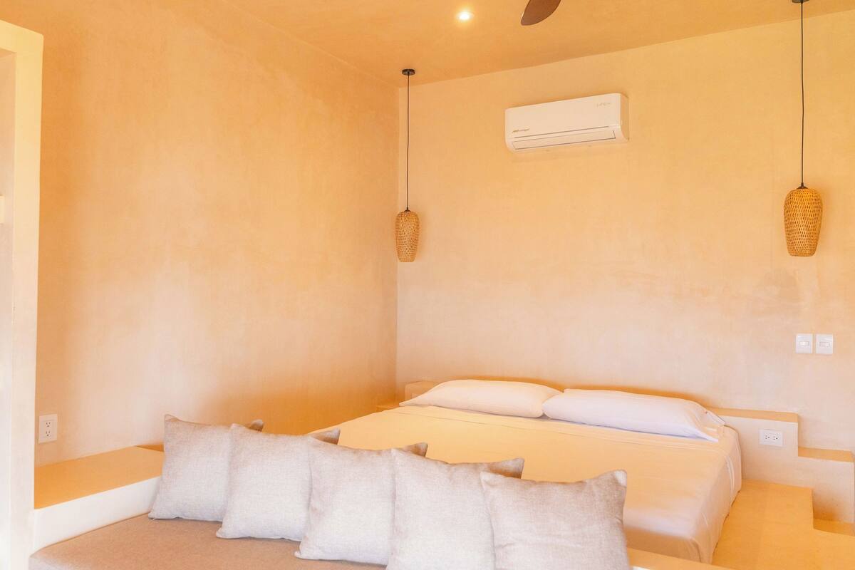 Un dormitorio con cama, almohadas, una estantería con un objeto verde, un equipo de aire acondicionado y una lámpara de fibra natural.