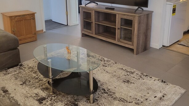 A living room with a glass coffee table, a flat-screen TV on a wooden stand, a grey sofa, and a patterned rug.
