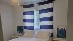 A bedroom with a bed, a nightstand, a ceiling fan, a window with striped curtains, and artwork on the nightstand.