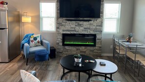 A modern living room with a stone fireplace, a flat-screen TV, a dining area with metal chairs, and a refrigerator. - 🌻Sunflower Cottage🌻Breathtaking Mountain Views & Fresh Air Retreat! (Flagstaff)