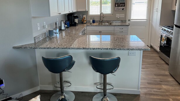 A modern kitchen with white cabinets, granite countertops, a stainless steel sink, and a built-in oven. There are two bar stools and a window with blinds. - 🌻Sunflower Cottage🌻Breathtaking Mountain Views & Fresh Air Retreat! (Flagstaff)