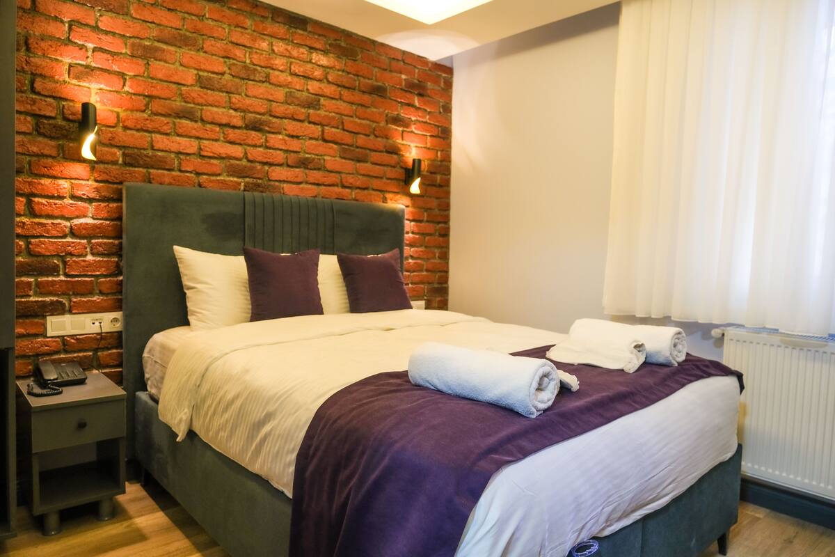 A hotel room with a brick wall, a green upholstered headboard, a white bedspread, and a purple throw blanket.