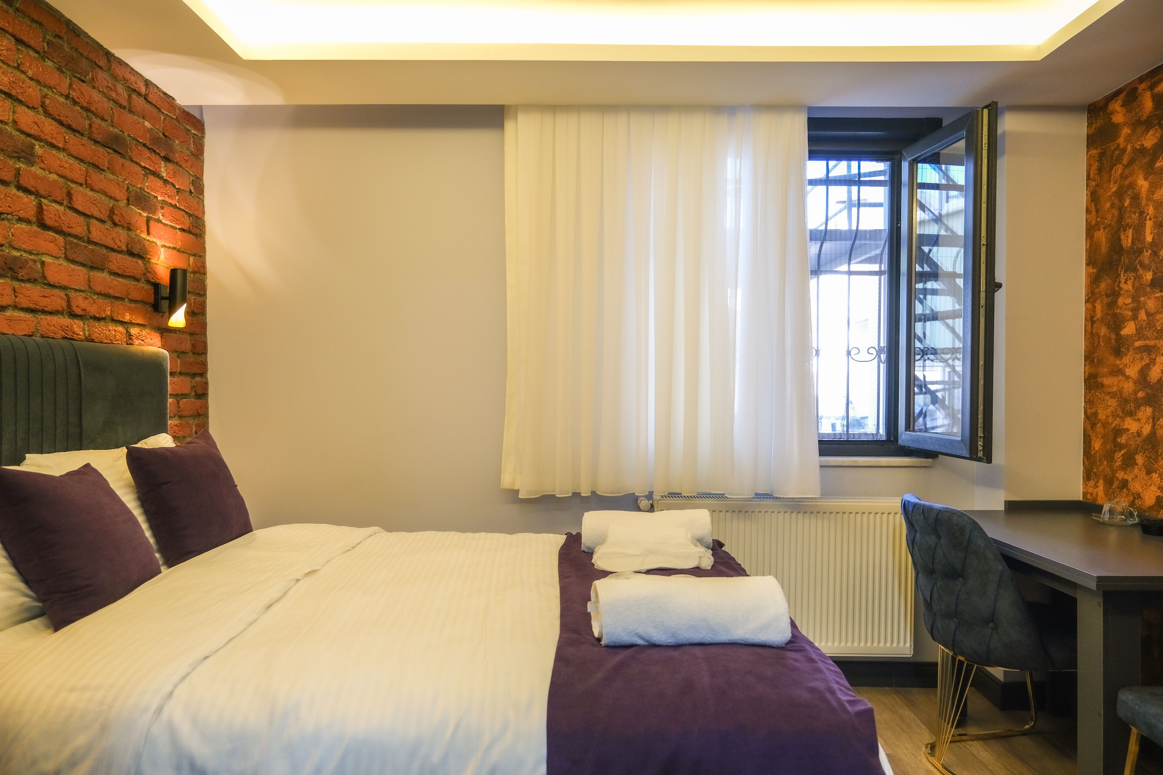 A hotel room with a bed, a desk, a chair, a window with curtains, and a brick wall.