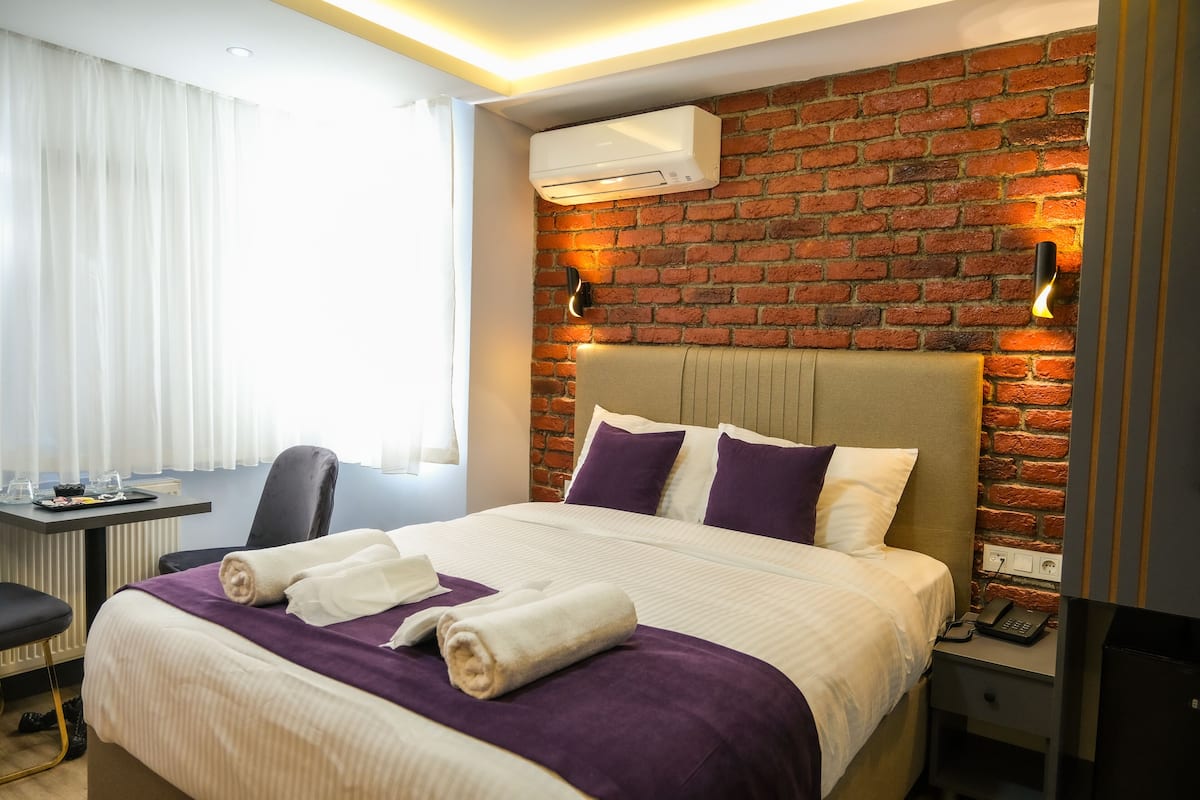 A hotel room with a brick wall, a bed with white and purple bedding, a desk with a chair, and an air conditioning unit.