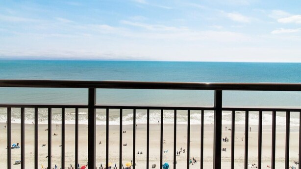 Une vue sur la plage depuis un balcon doté d’une balustrade, avec un ciel dégagé et une mer calme.