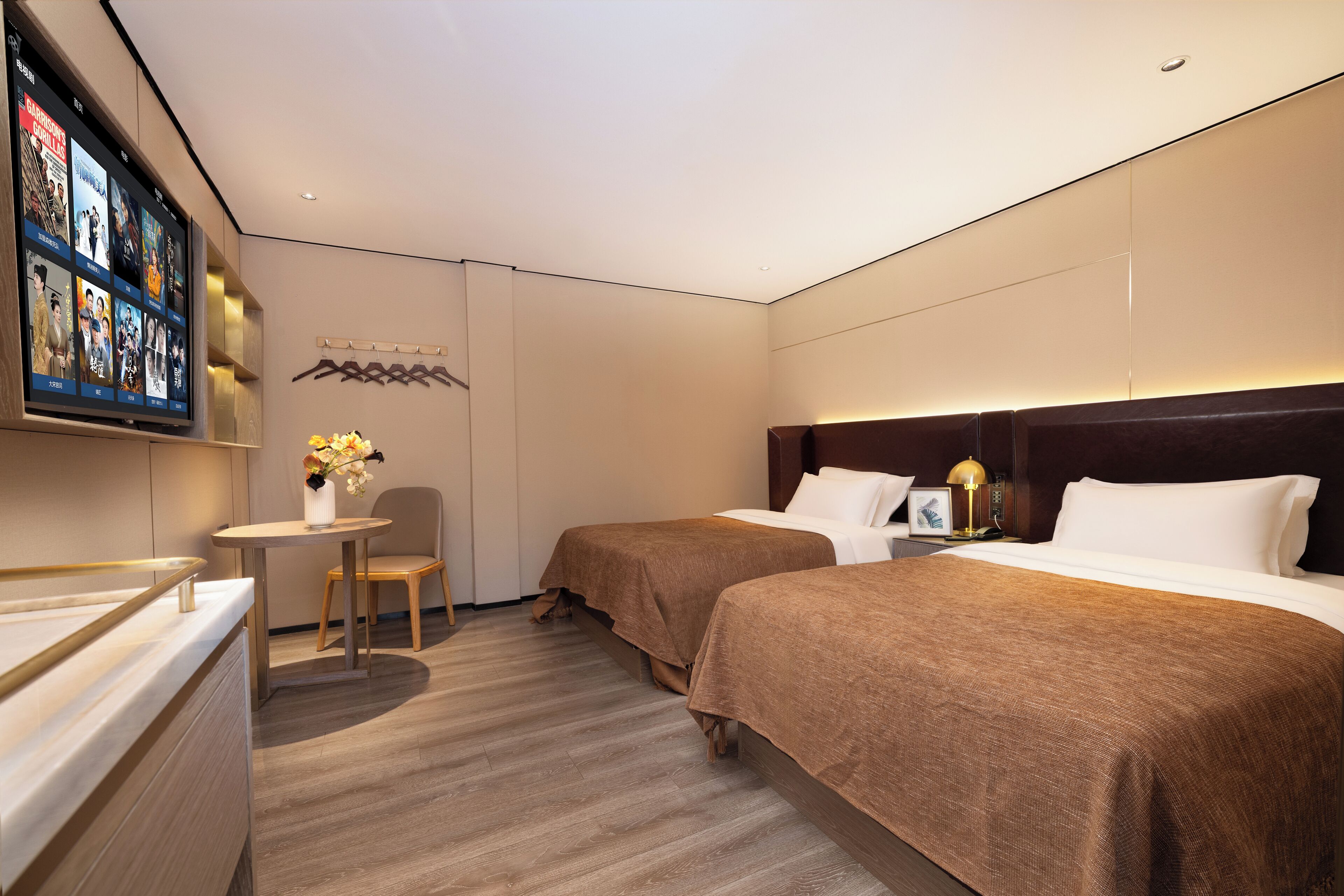 A hotel room with two beds, a small table with a vase of flowers, a television mounted on the wall, and a coat rack.