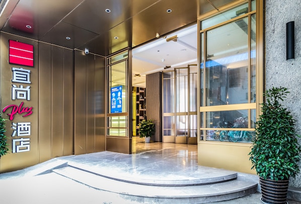 A hotel entrance with a modern design, featuring a glass door, a digital signboard, and a potted plant.