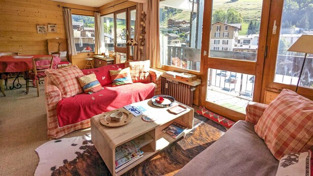 Ein gemütliches Blockhaus mit Holzvertäfelung, einem roten Sofa, einem kleinen Tisch mit einem Hut und Blick auf ein Dorf durch große Fenster.