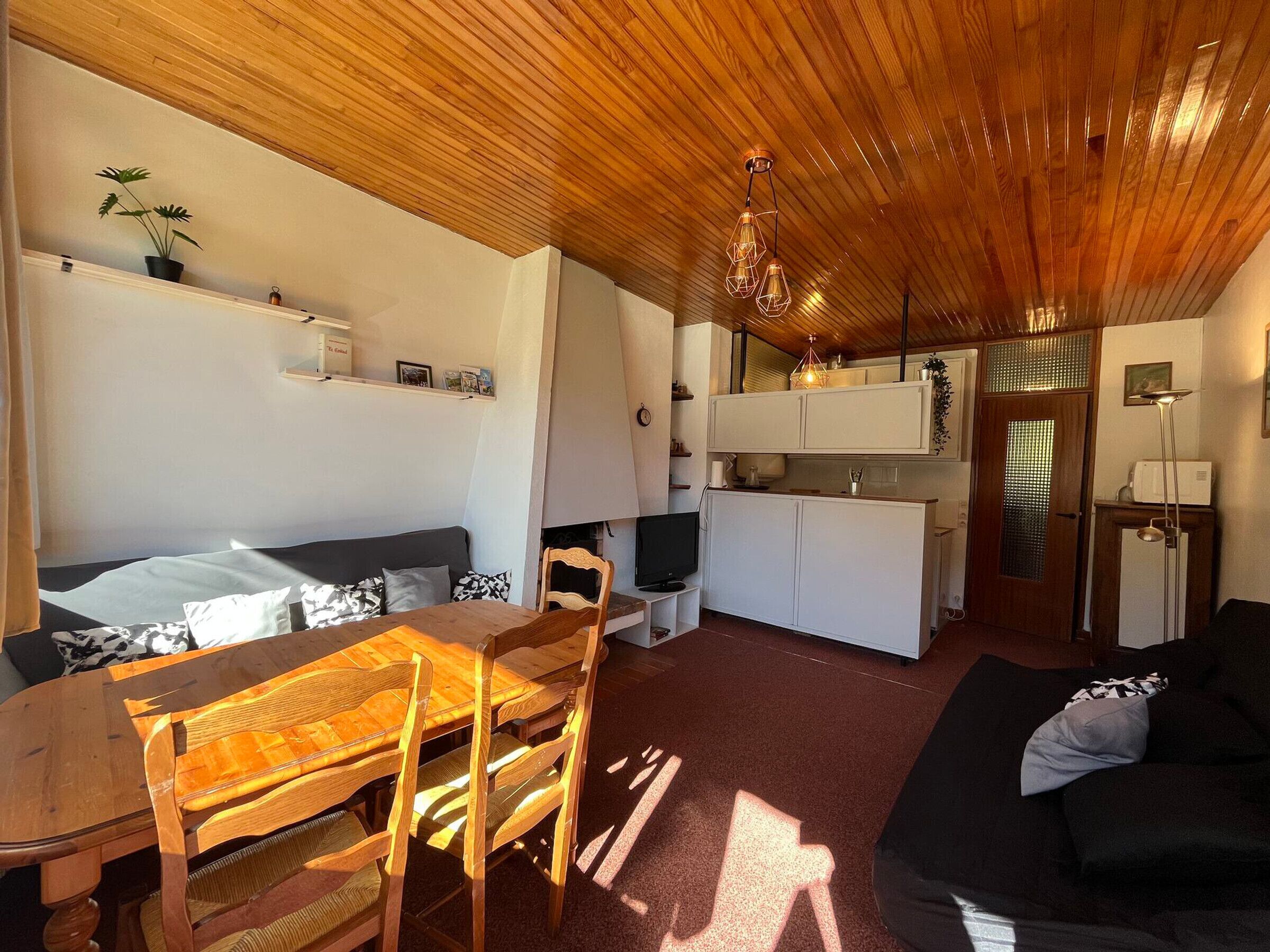 Un espacio habitable compacto con techo de madera, zona de comedor con mesa y sillas, sofá negro, kitchenette y televisor pequeño.