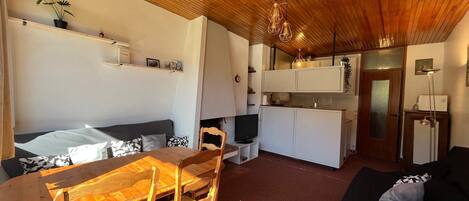 Un espacio habitable compacto con techo de madera, zona de comedor con mesa y sillas, sofá negro, kitchenette y televisor pequeño.