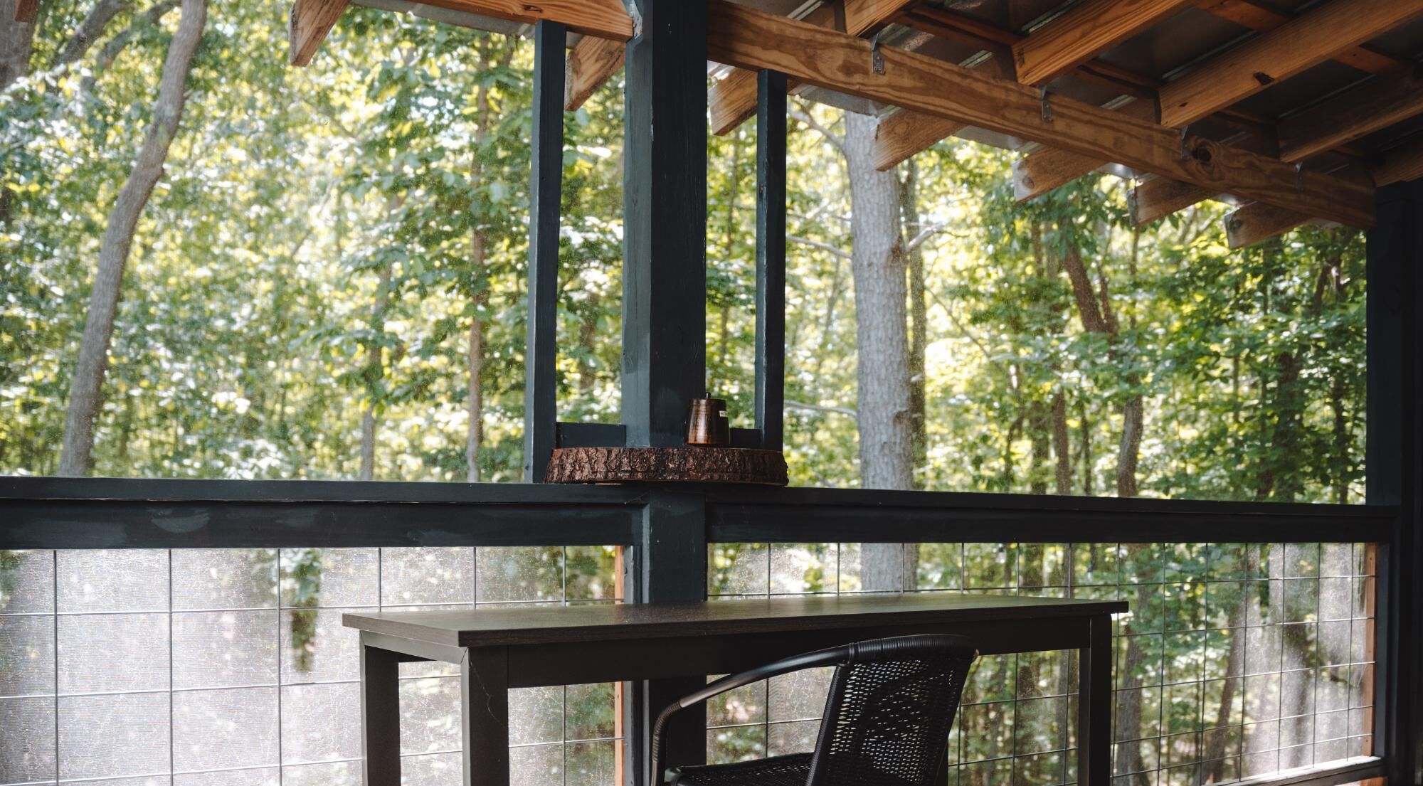 Un espacio al aire libre techado con una mesa y una silla, techo de madera y vista a árboles a través de una ventana.