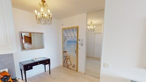 Une chambre d’hôtel avec un plancher en bois, un bureau avec des tiroirs, une chaise, un lit, un miroir, un lustre et une porte coulissante donnant sur une autre pièce.