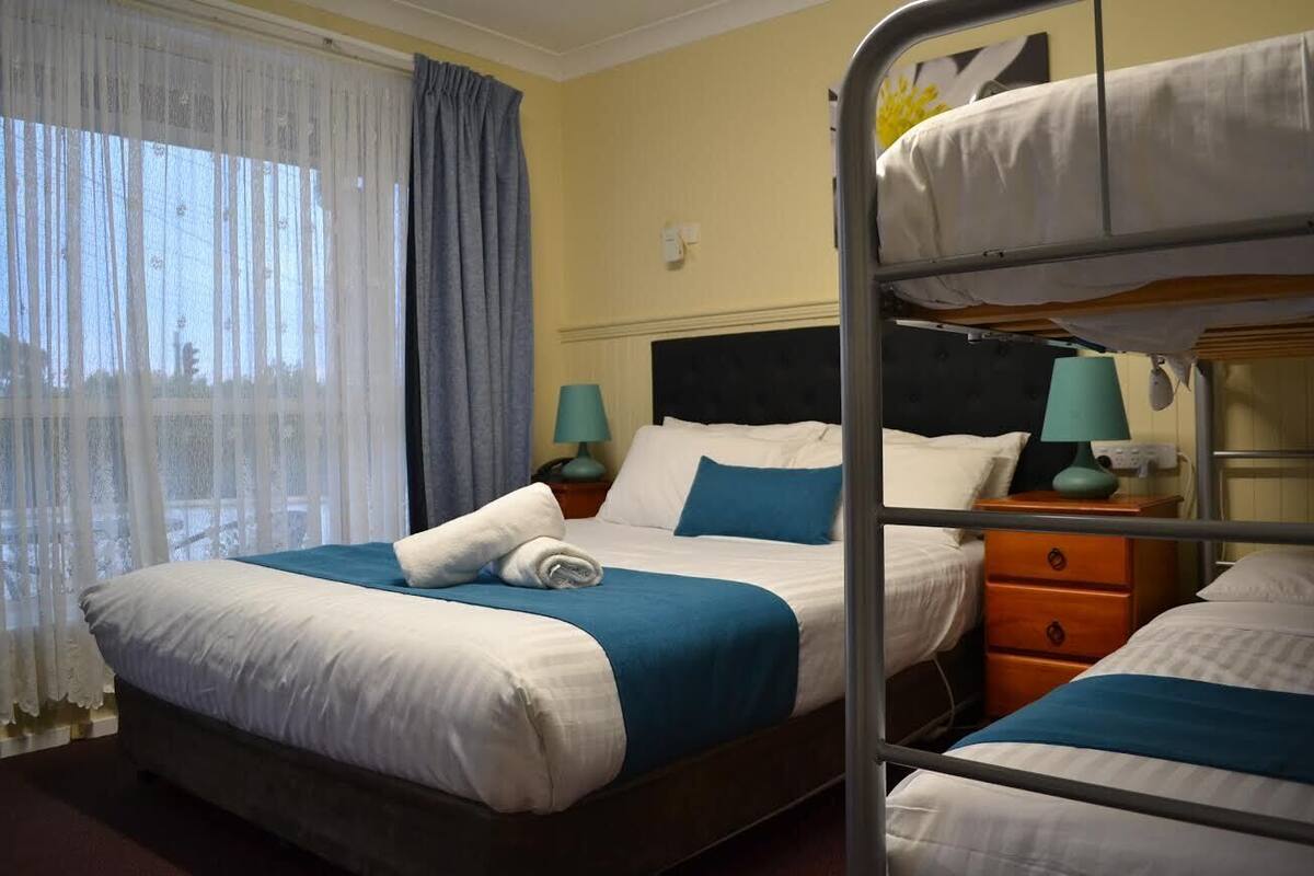 Una habitación de hotel con una litera, una cama individual y una mesita de noche con lámpara.
