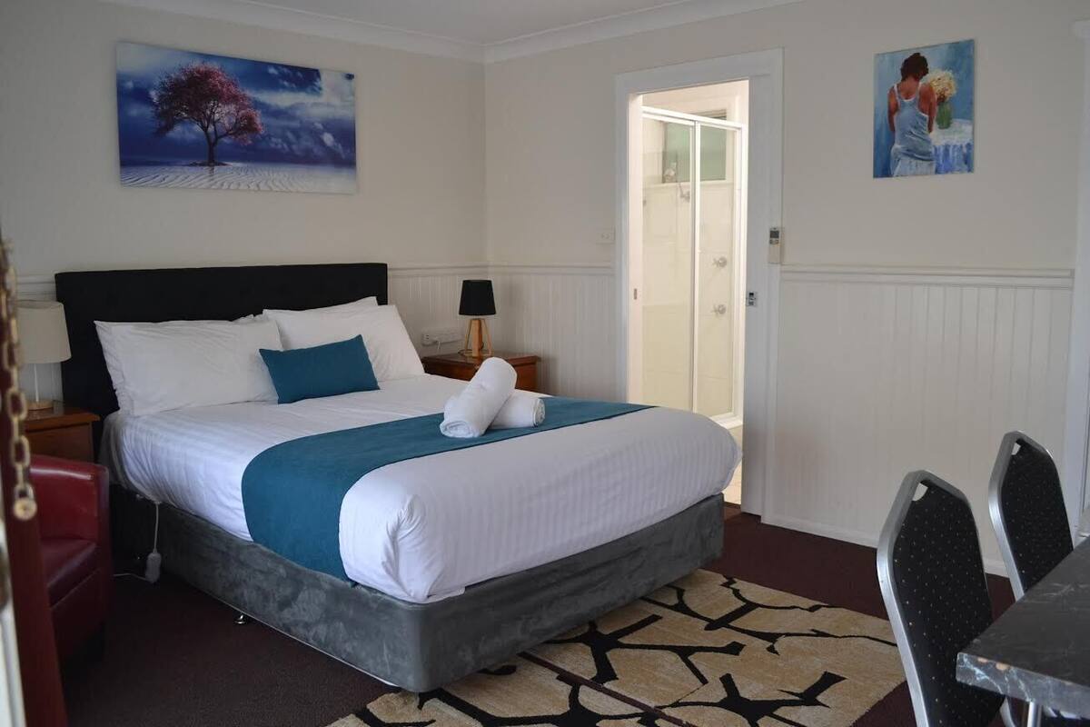 Habitación de hotel ordenada con una cama grande, una mesita de noche con lámpara, una silla y una mesa de comedor con sillas.