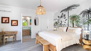 Een slaapkamer met een bed, een bankje, een hanglamp, een muur met een tropische plantenmuur en een zichtbare badkamer door een open deur.