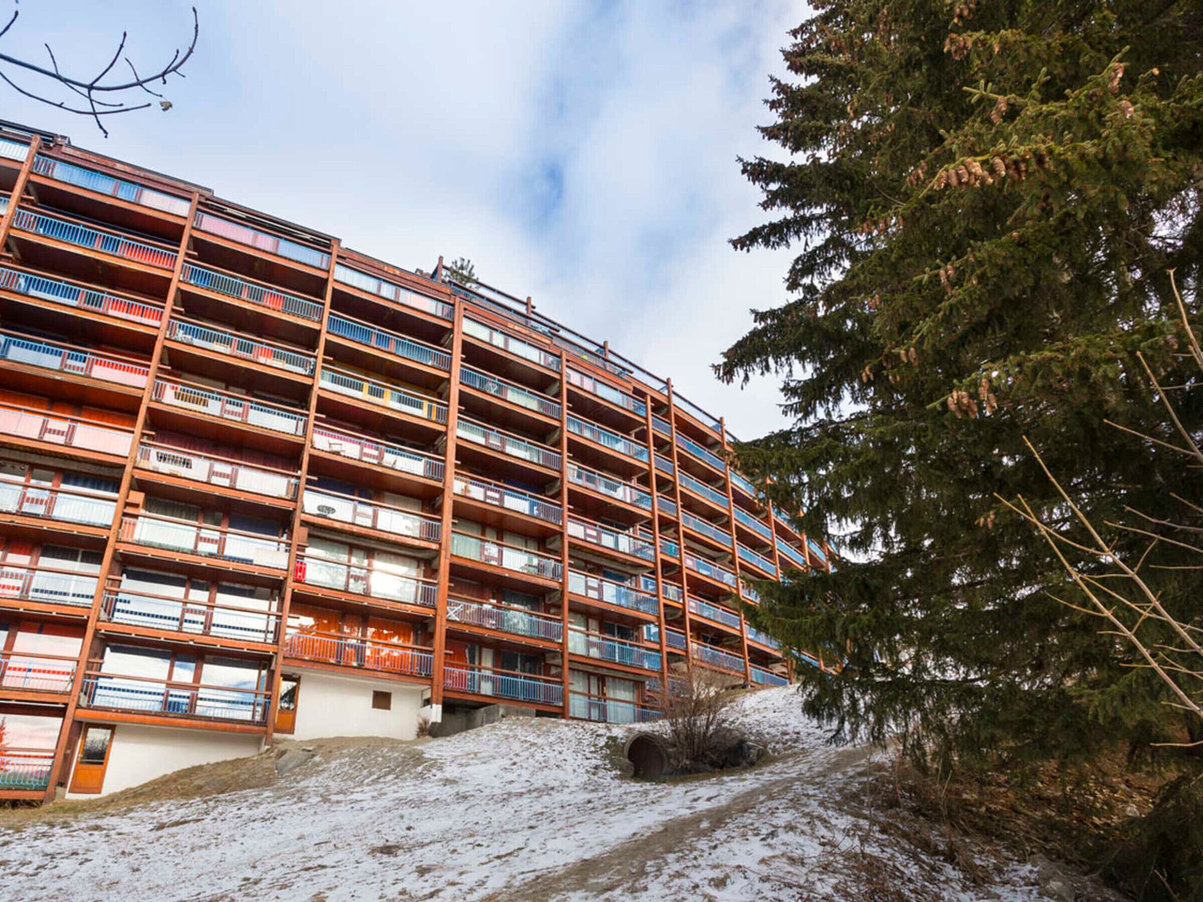 A multi-story hotel with balconies, a ski slope, and a large evergreen tree.