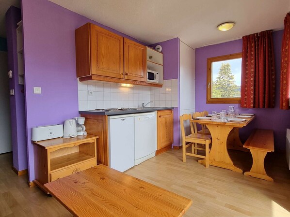 Cocina compacta con armarios de madera, microondas, cafetera y comedor con mesa y sillas de madera.