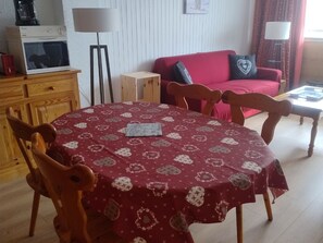 A dining area with a round table covered in a patterned tablecloth, wooden chairs, a red sofa, a small wooden cabinet, and a standing lamp. - Spacious central studio near gondola, sunny balcony (Châtel)