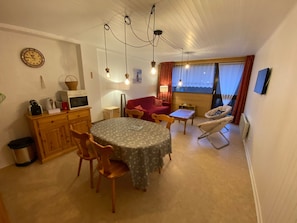 Ein kompakter Wohnraum mit Esstisch, StĂŒhlen, einem roten Sofa, einem Babywippstuhl und einem Fenster mit VorhĂ€ngen.