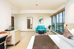 A modern hotel room with a bed, a sofa, a coffee table, a desk, and a large window offering a city view. - Presidency Airport Hotel (Aluva)