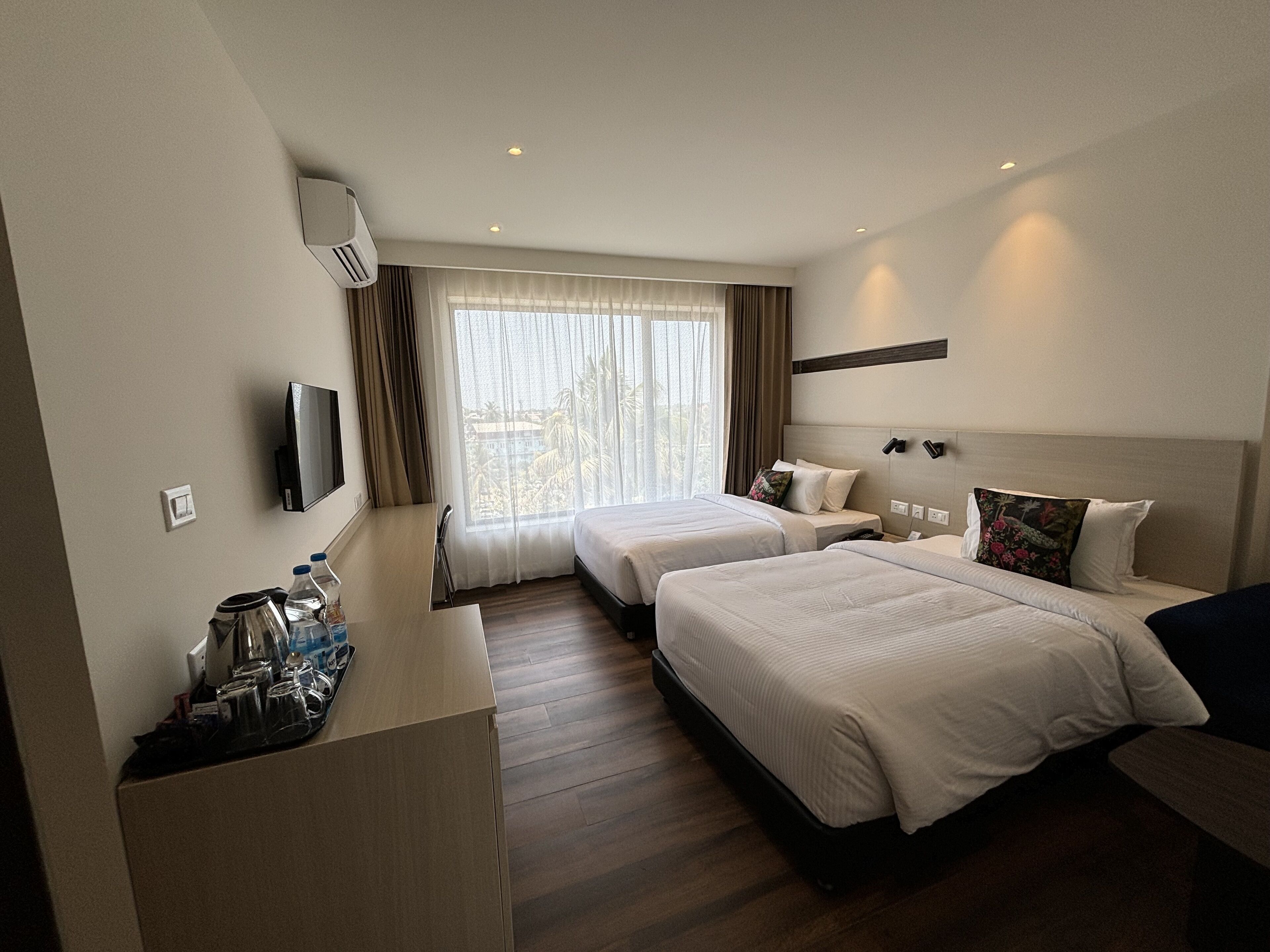 A hotel room with two beds, a TV, a desk with a coffee maker, and a window with curtains.