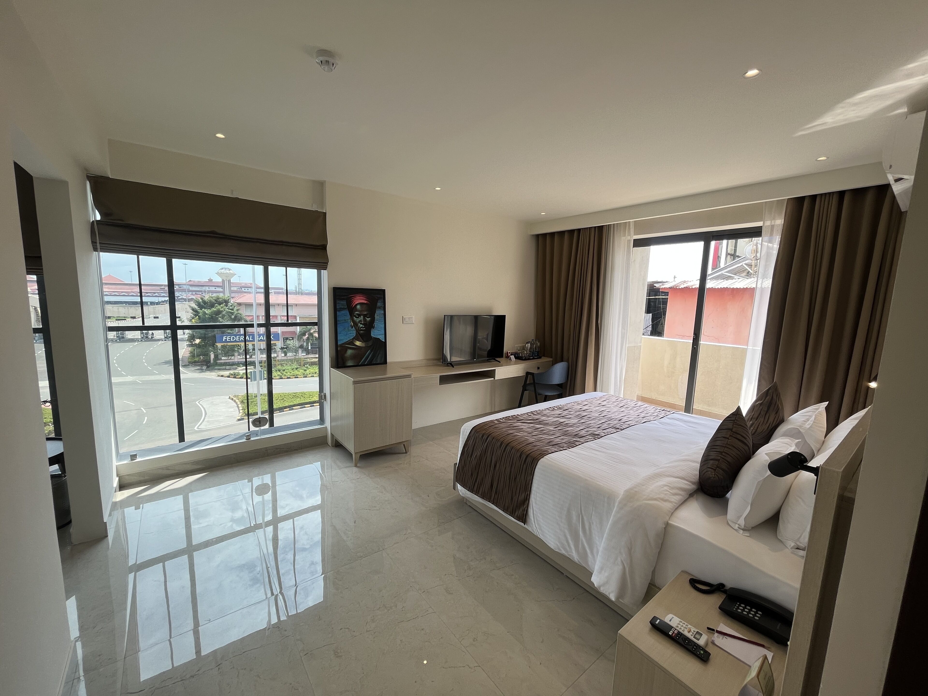 A hotel room with a large bed, a desk with a TV, a balcony with a view of the road, and a painting on the wall.