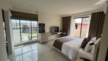 A hotel room with a large bed, a desk with a TV, a balcony with a view of the road, and a painting on the wall.