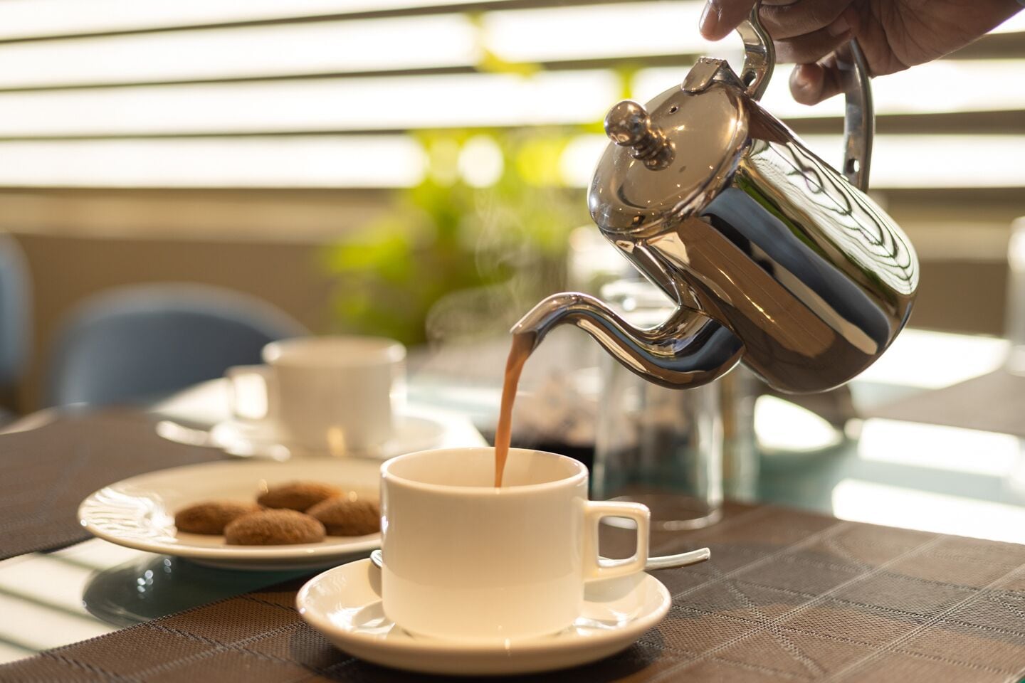 A hand pouring coffee into a cup on a table with a plate of cookies and a teapot.