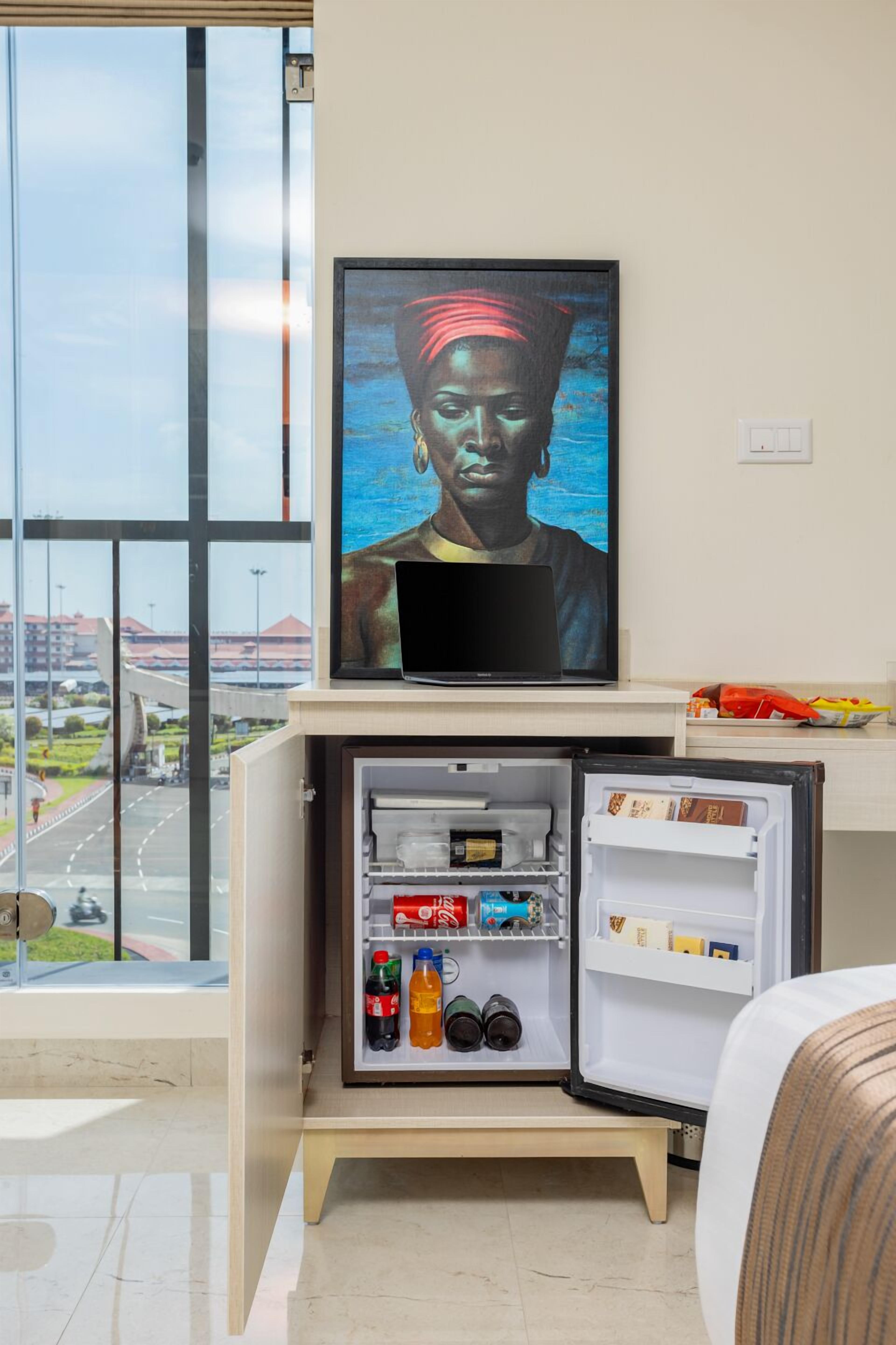 Ein kleines Zimmer mit einem eingebauten Minikühlschrank, gefüllt mit Getränken, einem darüber hängenden Fernseher und Blick auf eine Straße und Gebäude durch große Fenster.