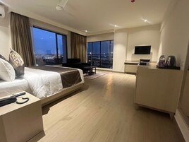 Una habitación de hotel moderna con una cama grande, un escritorio, una silla, un televisor y vistas de la ciudad al anochecer.