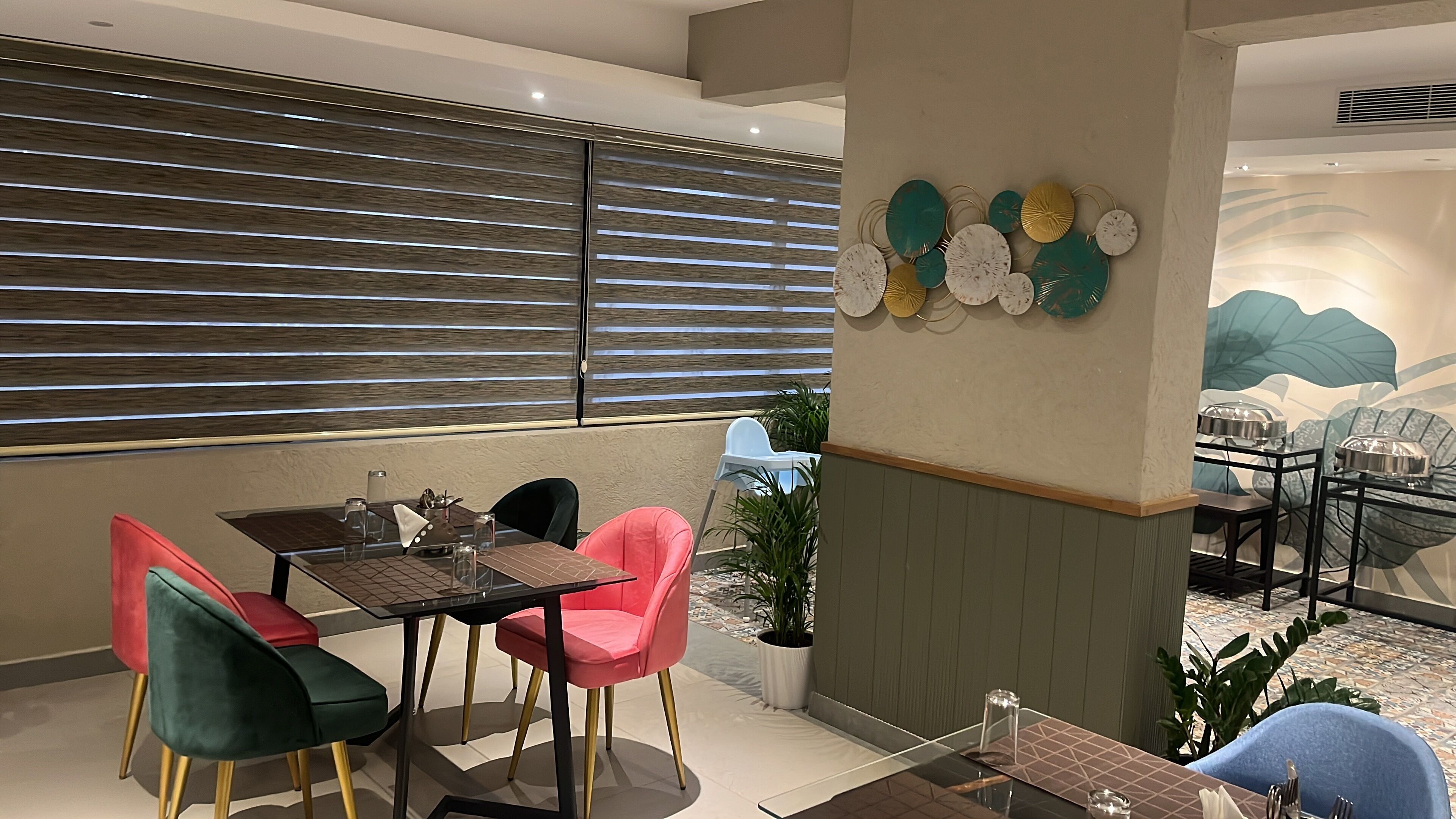 A dining area with a table set for two, surrounded by colorful chairs, a decorative wall with circular patterns, and a glimpse of a kitchen area in the background.