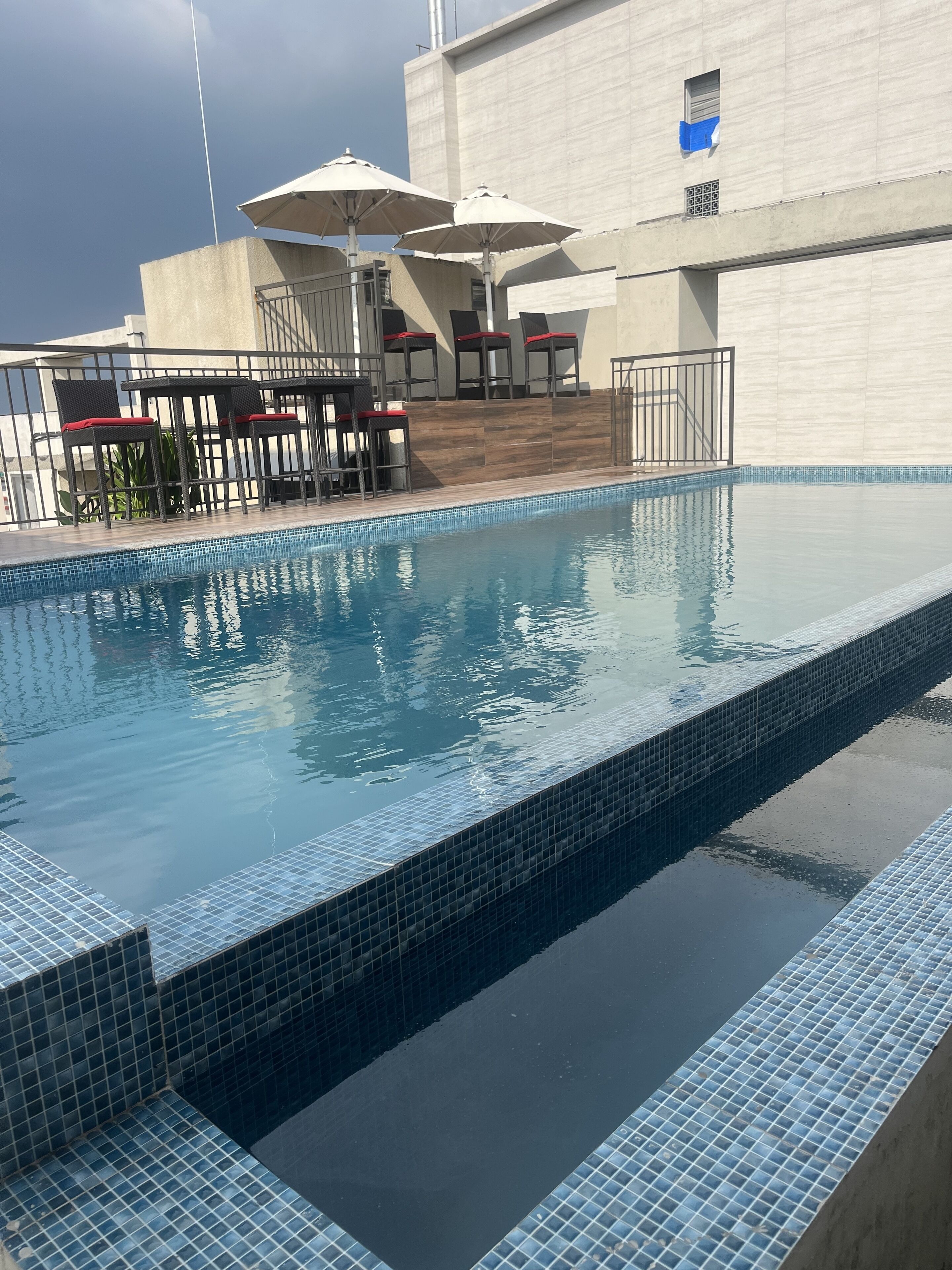 A swimming pool with tiled edges, outdoor seating with umbrellas, and a building in the background.