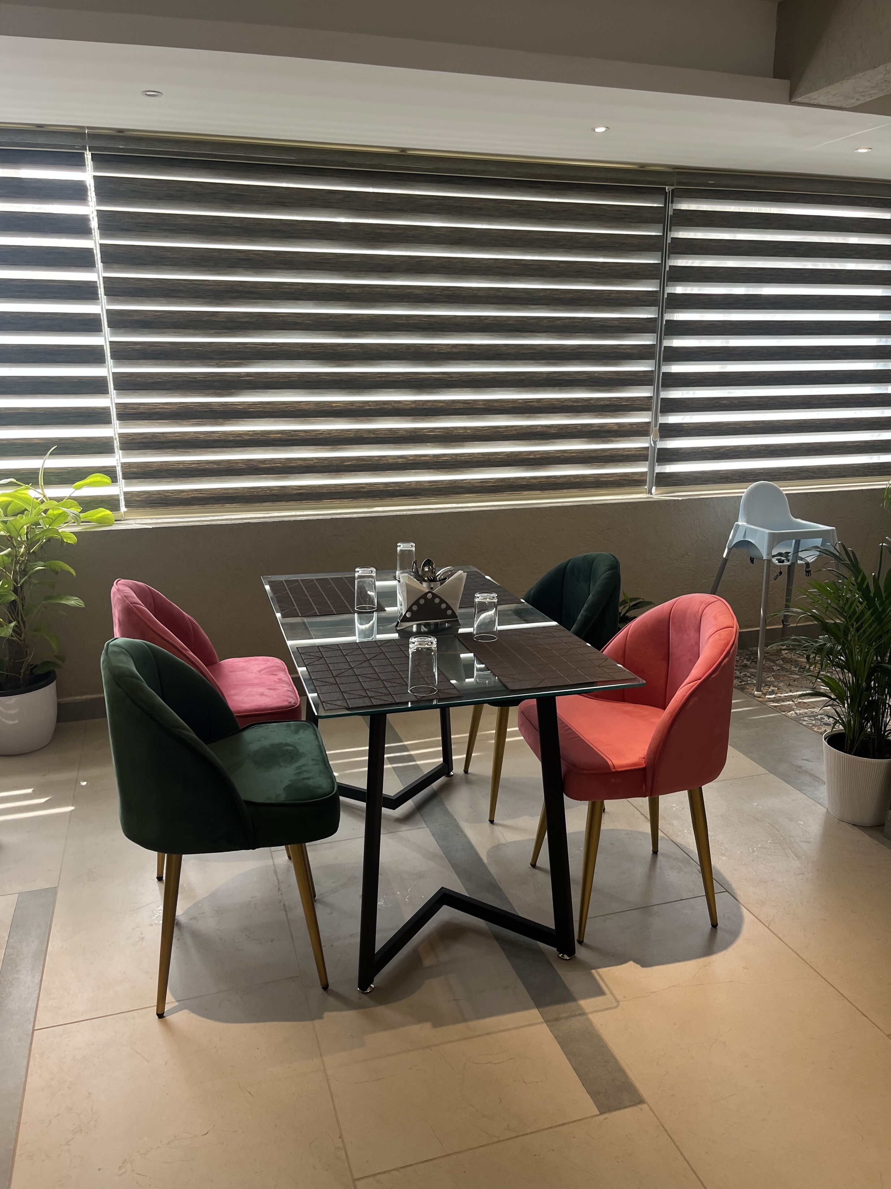 Un comedor con una mesa de vidrio y cuatro sillas, dos verdes y dos rojas, con vistas a un edificio a través de amplias ventanas.