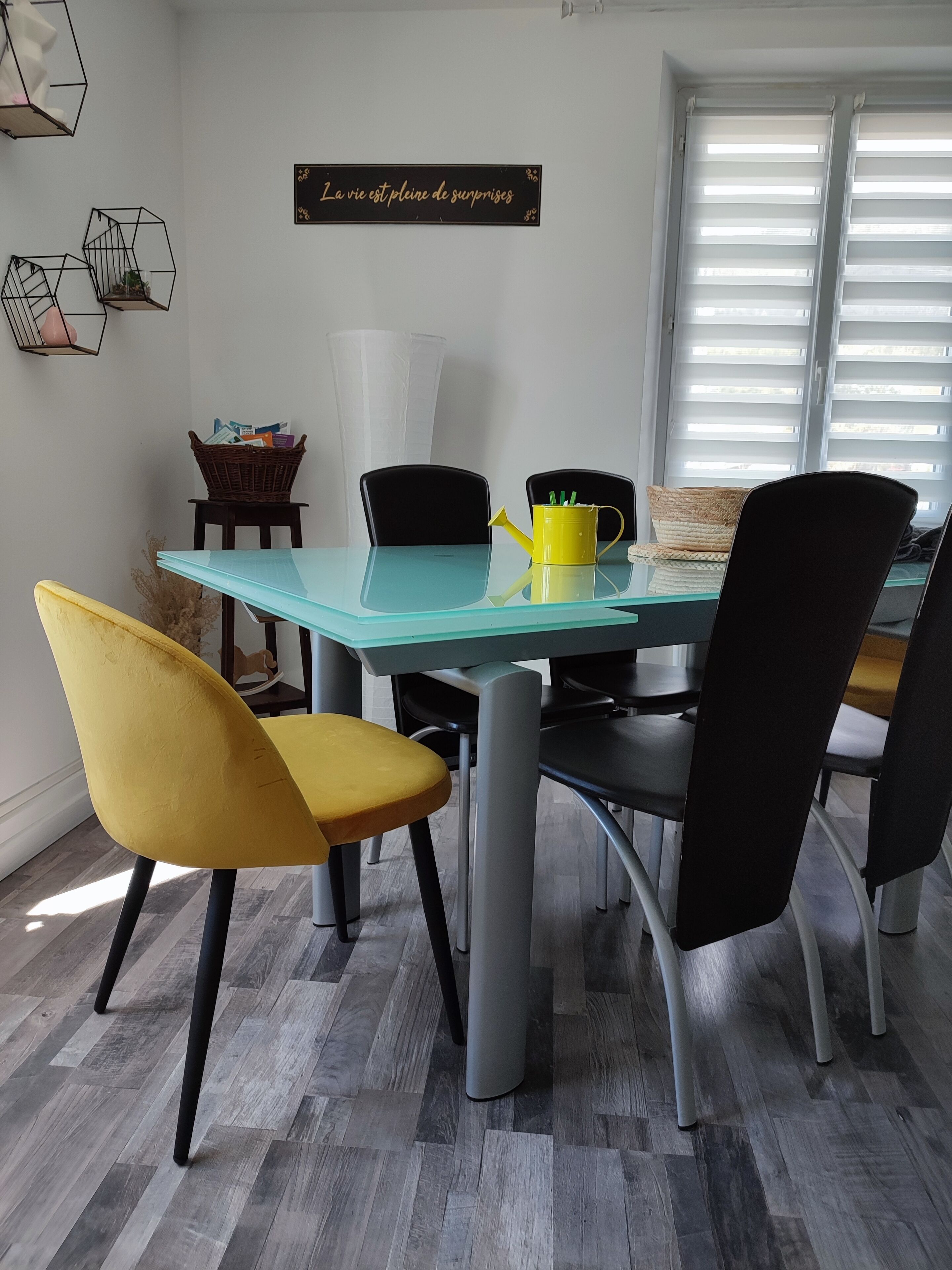 A dining area with a glass table, black and yellow chairs, a yellow watering can, and a wall sign.