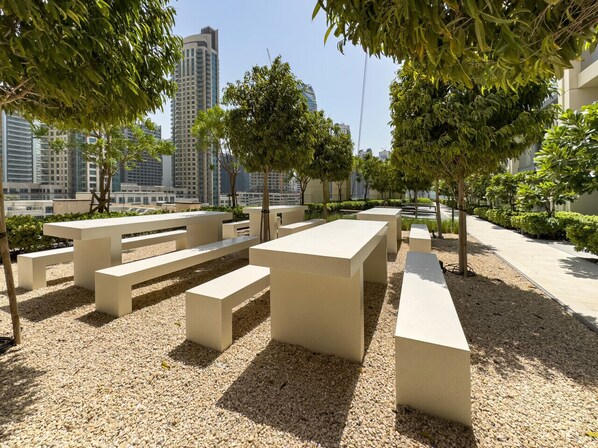 An outdoor seating area with concrete benches and tables, surrounded by trees and a cityscape in the background.