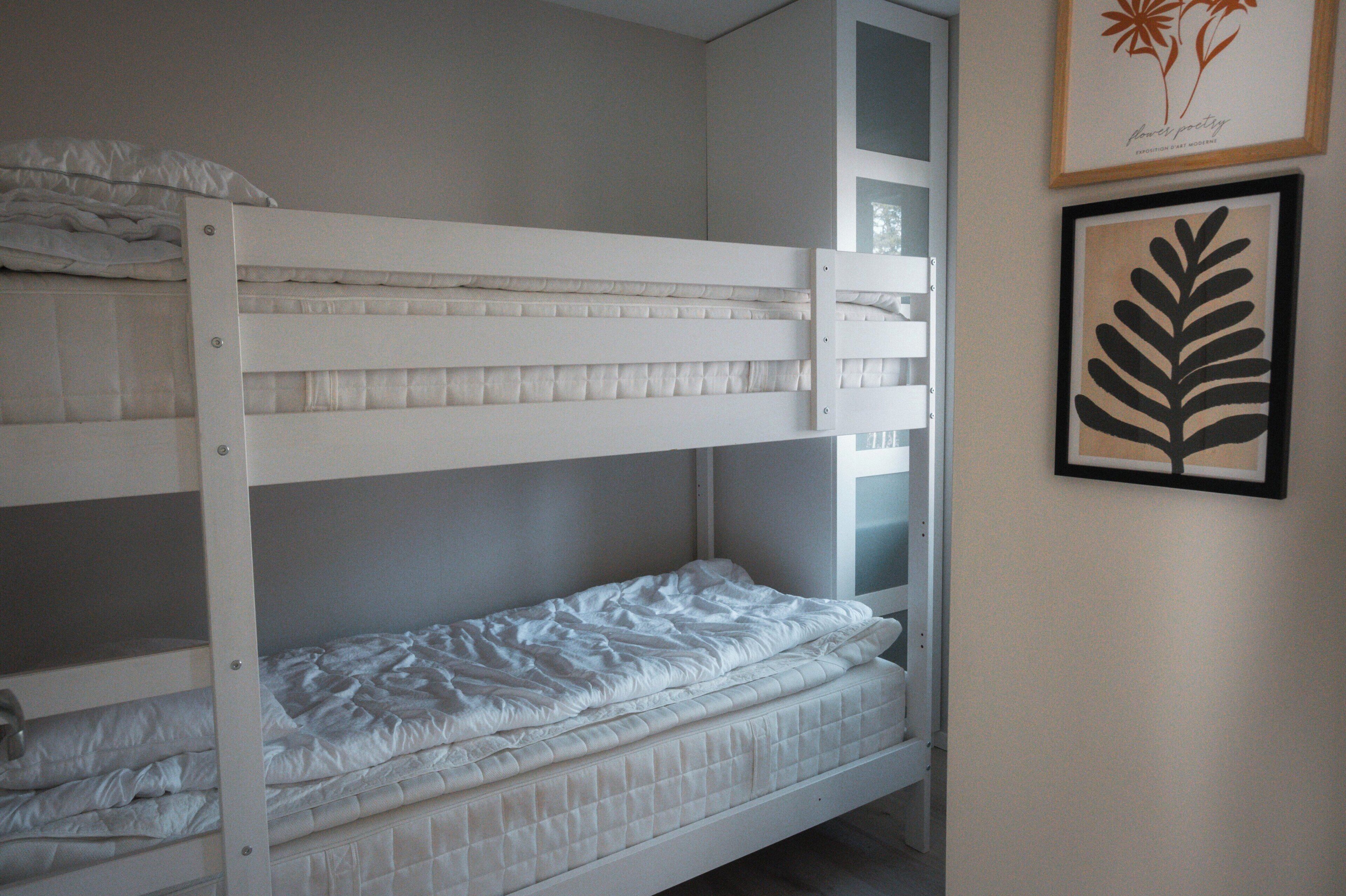 Ein Stockbett mit Matratze und Kissen, ein gerahmtes Kunstwerk an der Wand und ein Fenster mit Jalousien.