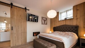 Une chambre avec un grand lit, un banc, une cheminée, des murs en bois et un luminaire suspendu.