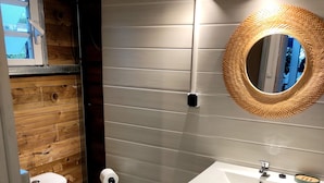 A modern bathroom with a wooden floor, a white toilet, a white sink, and a mirror with a woven frame. - Chambre Privée Chez Jo, en Bordure de Savane (Saint Paul)