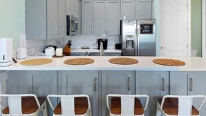 Een moderne keuken met grijze kastjes, een roestvrijstalen koelkast en een koffiezetapparaat. Er staan vier barkrukken met houten zitplaatsen en geweven onderzetters.